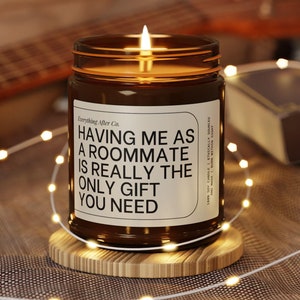 May include: A brown glass candle with a white label that reads "Having me as a roommate is really the only gift you need". The candle is lit and sitting on a wooden coaster.