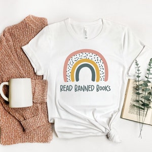 May include: White t-shirt with a rainbow graphic and the text "Read Banned Books". The rainbow is pink, yellow, and blue with black polka dots. The t-shirt is on a white surface with a brown sweater and a book.