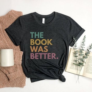 May include: A dark gray t-shirt with the text "THE BOOK WAS BETTER." in colorful letters. The shirt is on a white surface with a book and a sprig of greenery.