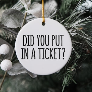 May include: White ceramic Christmas ornament with black text that reads "Did you put in a ticket?" hanging on a green Christmas tree.
