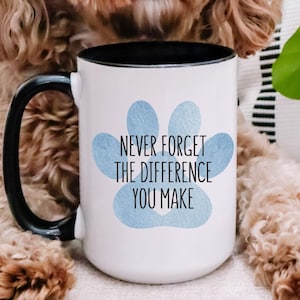 May include: A white ceramic mug with a black handle and rim, featuring a blue paw print design and the text "NEVER FORGET THE DIFFERENCE YOU MAKE." A fluffy brown dog is in the background. The mug is approximately 4.7 inches tall.