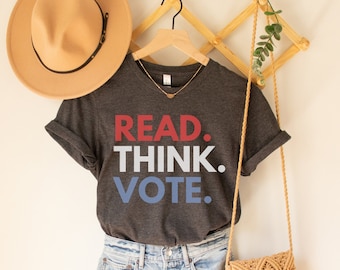 Op de afbeelding: Een donkergrijs t-shirt met de woorden "READ. THINK. VOTE." in rood, wit en blauw. Het shirt wordt gecombineerd met een spijkerbroek, een beige hoed en een geweven crossbodytas. Een houten hanger en wanddecoratie maken de presentatie compleet.