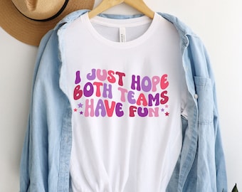 May include: White t-shirt with the text "I just hope both teams have fun" in pink, purple, and blue letters with stars on either side of the text.