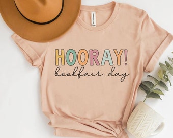 May include: A light pink t-shirt with the text "HOORAY! bookfair day" in colorful letters. The shirt is folded and displayed on a white surface with a brown hat and a white mug.