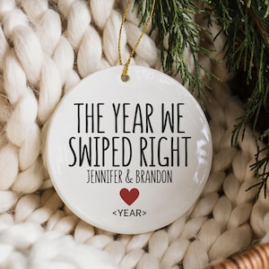 May include: A white ceramic Christmas ornament with the text "THE YEAR WE SWIPED RIGHT" in black, the names "Jennifer & Brandon" and a red heart. It hangs by a gold string, set against a white knitted background, with greenery.