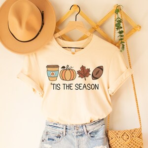 May include: A beige t-shirt with a graphic of a coffee cup, pumpkin, leaf, and football with the text 'Tis the season'. The shirt is hanging on a wooden hanger with a brown hat and a woven bag.