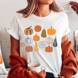May include: White t-shirt with a colorful pumpkin design. The shirt has a crew neck and short sleeves. The design features a variety of pumpkins in different colors and shapes. The text "Pick Your Own Locally Grown Pumpkin Patch" is on a pillow in the background.