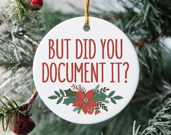 May include: A white ceramic Christmas ornament with the text "BUT DID YOU DOCUMENT IT?" in red. The ornament features a floral design with red poinsettias and green leaves. It hangs from a gold string.