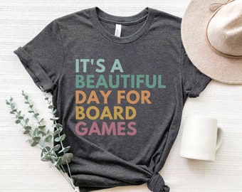 Könnte beinhalten: Ein dunkelgraues T-Shirt mit dem Text "It's a beautiful day for board games" in bunten Buchstaben.
