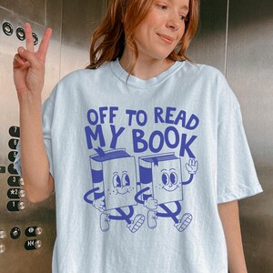 May include: Light blue t-shirt with the text "OFF TO READ MY BOOK" in blue. The graphic features two cartoon books with smiling faces and legs. The shirt is worn by a person.
