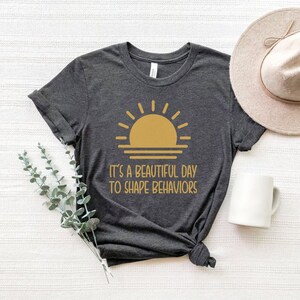 May include: A dark gray t-shirt with a yellow sun graphic and the text "It's a beautiful day to shape behaviors".