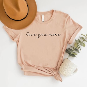 May include: A light pink t-shirt with the text "love you more" written in black cursive script.
