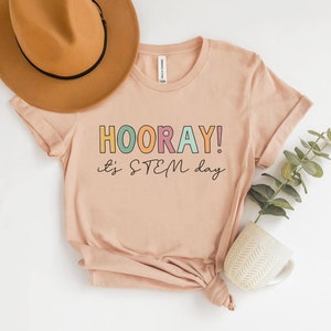 Pode incluir: Uma camiseta rosa claro com o texto "HOORAY! It's STEM day" em letras coloridas.