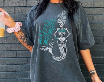 May include: A dark gray oversized t-shirt with a skeleton mermaid graphic in white and teal. The mermaid is wearing a seashell bra and has long flowing hair. The text "Life is tough, my darling, but so are you" is tattooed on the wearer's leg.