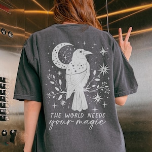 May include: Black t-shirt with a white graphic of a crow perched on a branch with a crescent moon and stars. The text "The world needs your magic" is printed below the crow.