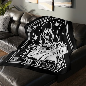 May include: Black and white throw blanket with a skeleton reading a book. The blanket features a tarot card design with the text "The Reader".