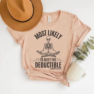 May include: A pale pink t-shirt featuring the words "MOST LIKELY TO MEET THE DEDUCTIBLE" and a meditating skeleton illustration. The hashtag #CHRONICILLNESS is below. A brown hat and a white mug with a plant are in the background.