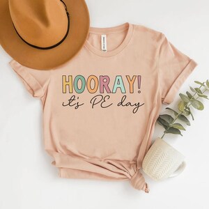May include: A peach-colored t-shirt with the text "HOORAY! it's PE day" in colorful block letters. A tan hat with a black band sits above the shirt. A small white ceramic pot with a plant is to the right of the shirt.
