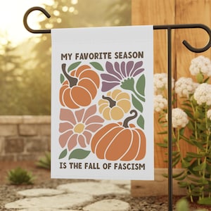 May include: A white garden flag with the text "MY FAVORITE SEASON IS THE FALL OF FASCISM" and a fall-themed design of pumpkins and flowers in orange, yellow, purple, and green. The flag is displayed on a black metal garden stake.