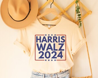 May include: A beige t-shirt with a red and blue rectangle graphic that says "HARRIS WALZ 2024" with stars above and below the text. The shirt is hanging on a wooden hanger.