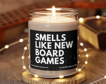Könnte beinhalten: Eine braune Kerze mit einem schwarzen Etikett, auf dem "Smells Like New Board Games" steht. Die Kerze steht auf einem Holzuntersetzer und ist von weißen Lichterketten umgeben.
