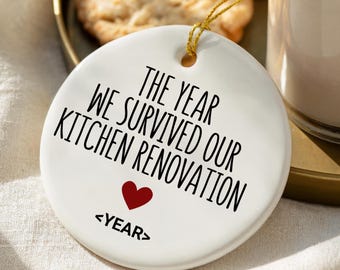 May include: A white ceramic ornament with gold string, featuring the text "THE YEAR WE SURVIVED OUR KITCHEN RENOVATION" in black, with a red heart and the word "<YEAR>" below. The ornament is on a gold tray with cookies and a drink.