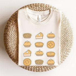 May include: A cream-colored t-shirt with a pattern of various pie designs in shades of yellow and orange. The pies are drawn in a simple, cartoon style.