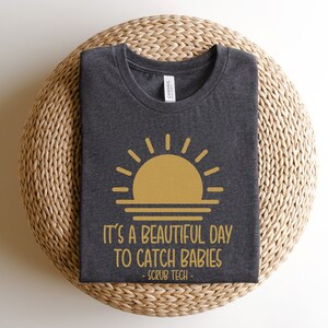 May include: A dark gray t-shirt with a gold sunset graphic and the text "It's a beautiful day to catch babies - Scrub Tech -" printed on the front.