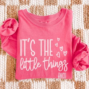 May include: A pink long-sleeved shirt with the text "IT'S THE little things" in white script, along with three small heart graphics. The hashtag #NICU is below the text. The shirt is folded on a woven, neutral-toned surface.