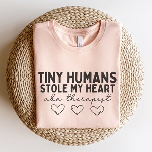 May include: A pink t-shirt with the text "Tiny Humans Stole My Heart aka therapist" and three small hearts.
