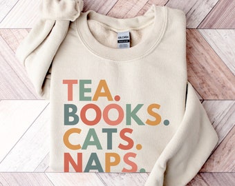 Peut inclure: Un sweat-shirt crème avec le texte "TEA. BOOKS. CATS. NAPS." en lettres majuscules colorées. Les lettres sont dans des tons d'orange, de vert, de jaune et de rose.