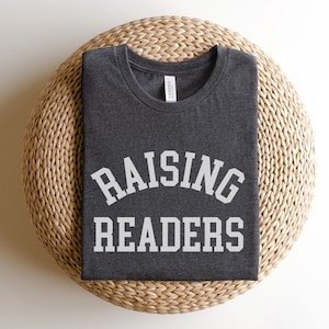 May include: A dark gray t-shirt with the text "RAISING READERS" in white letters. The shirt is folded and sitting on a woven straw mat.