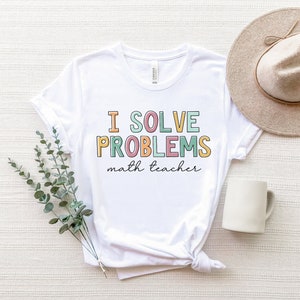 May include: White t-shirt with the text "I solve problems math teacher" in colourful letters. The shirt is laid flat on a white surface with a hat and a mug.