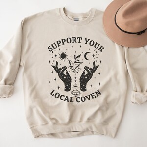 Puede incluir: Una sudadera beige con el texto "Support Your Local Coven" impreso en negro. El dise&ntilde;o incluye un par de manos sosteniendo una planta con una luna, un sol y estrellas.
