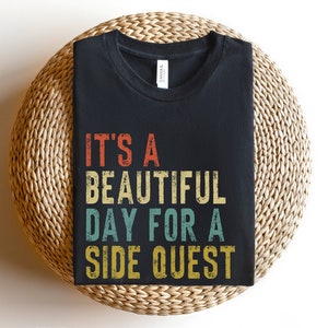 May include: Black t-shirt with a vintage-style graphic that reads "It's a beautiful day for a side quest".
