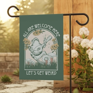 May include: A green garden flag with a whimsical design. The flag features a frog playing a stringed instrument, surrounded by sunflowers and mushrooms. Text on the flag reads "ALL ARE WELCOME HERE" and "LET'S GET WEIRD". The flag is displayed on a black metal stand.