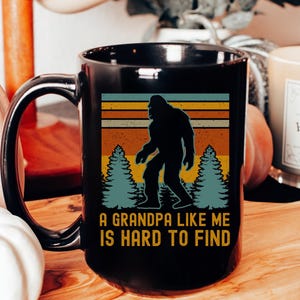May include: Black coffee mug with a Bigfoot silhouette design against a retro sunset background. The mug features the text "A Grandpa Like Me Is Hard To Find". The mug is displayed on a wooden serving board.