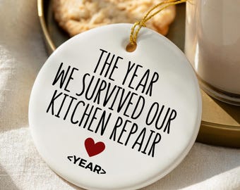 May include: A white ceramic ornament with gold string, featuring the text "THE YEAR WE SURVIVED OUR KITCHEN REPAIR" in black, with a red heart and the word "<YEAR>". The ornament is on a gold tray with cookies and a glass of dessert.