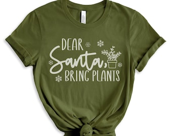 May include: Olive green t-shirt with the text "DEAR Santa, BRING PLANTS" in white script. The design includes snowflakes and a potted plant graphic. The shirt is knotted at the bottom.