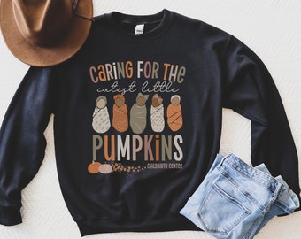 Puede incluir: Sudadera negra con el texto "Caring for the cutest little pumpkins" e ilustraciones de bebés envueltos. El diseño incluye calabazas y hojas de otoño. Un sombrero marrón y unos vaqueros azules también están en la imagen.