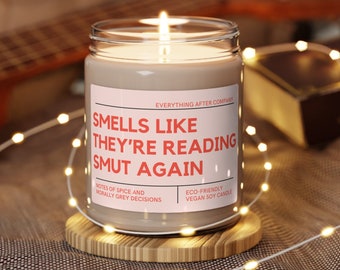 May include: A lit soy candle in a glass jar with the text "Smells Like They're Reading Smut Again." The candle sits on a wooden coaster, surrounded by fairy lights. The label also reads "Notes of Spice and Morally Grey Decisions."