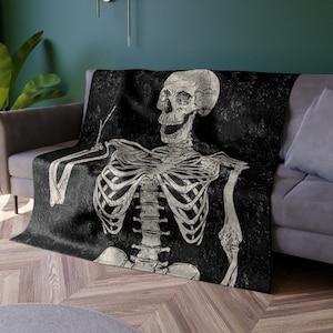 May include: A black and white throw blanket with a skeleton design. The skeleton is sitting with its arms crossed and has a distressed look. The blanket has a vintage, grunge aesthetic.
