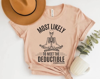May include: A light peach-colored t-shirt with the text "MOST LIKELY TO MEET THE DEDUCTIBLE" and a meditating skeleton graphic. The hashtag #CHRONICILLNESS is below the text. A brown hat and a white mug with a plant are in the background.