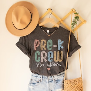 May include: A gray t-shirt with the text "Pre-K Crew" in colorful letters and "Mrs. Williams" below. The shirt is hanging on a wooden hanger with a brown hat and a straw bag.