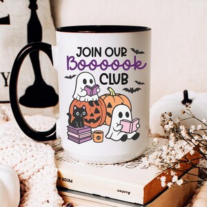 May include: White ceramic mug with a black handle and rim, featuring the text "JOIN OUR BOOOOOK CLUB" in purple. The mug has illustrations of ghosts, pumpkins, and a black cat. The mug is surrounded by white pumpkins and books, creating a Halloween theme.