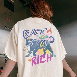 Pode incluir: Camiseta creme com um gr&aacute;fico nas costas. O gr&aacute;fico apresenta um tigre com o texto "EAT THE RICH" em roxo, azul e amarelo. O tigre &eacute; preto e amarelo com chamas e borboletas.