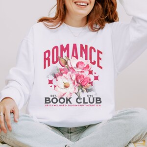 May include: White sweatshirt with the text "ROMANCE BOOK CLUB" in a maroon collegiate font. A floral graphic with pink and red flowers is centered below the text. The sweatshirt also has the text "EST. 1740 SPICY CLOSED DOORS SMUT EROTICA".