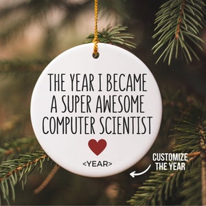May include: White ceramic Christmas bauble with the text "THE YEAR I BECAME A SUPER AWESOME COMPUTER SCIENTIST" and a red heart. The text "<YEAR>" is below the heart. The bauble is hanging on a green Christmas tree.