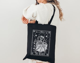 May include: Black tote bag with a white design of a skeleton reading a book. The design includes the text "The Reader" and celestial symbols.