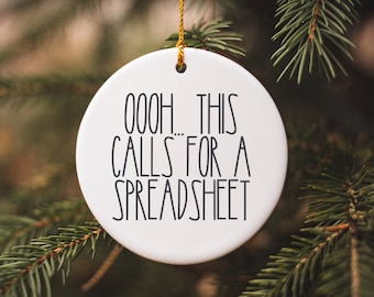 May include: White round Christmas ornament with black text that reads "OOOH... THIS CALLS FOR A SPREADSHEET". The ornament is hanging from a green pine tree branch.